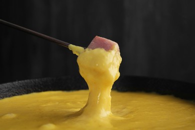 Photo of Dipping piece of ham into fondue pot with tasty melted cheese against dark gray background, closeup