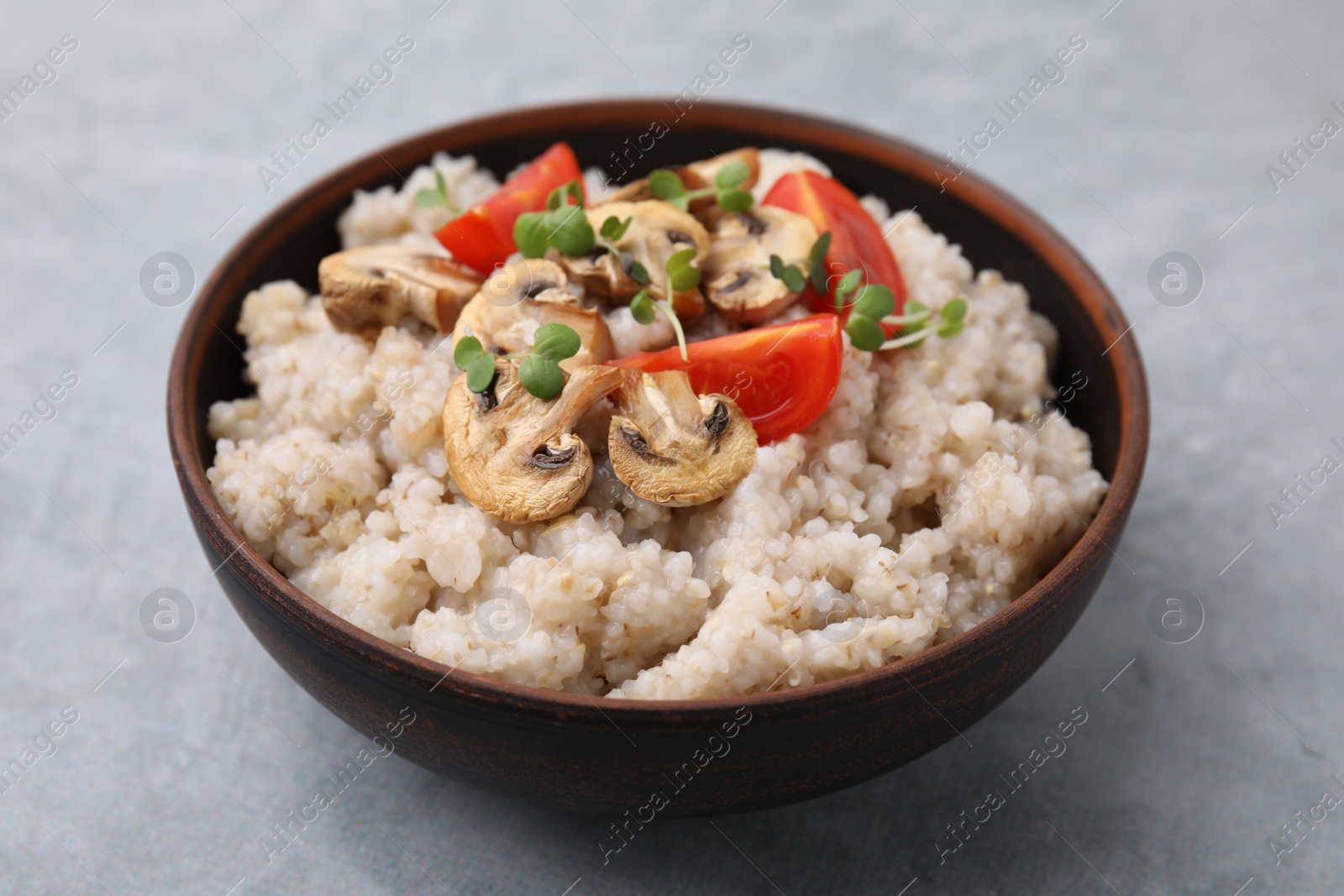 Photo of Delicious barley porridge with mushrooms, tomatoes and microgreens in bowl on gray table, closeup