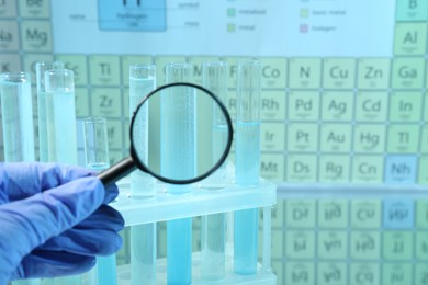 Photo of Laboratory assistant looking through magnifying glass on test tubes against periodic table of chemical elements, closeup. Space for text