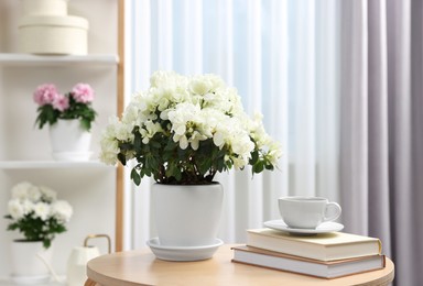 Photo of Beautiful azalea plant in flower pot, cup of coffee and books on wooden table in room