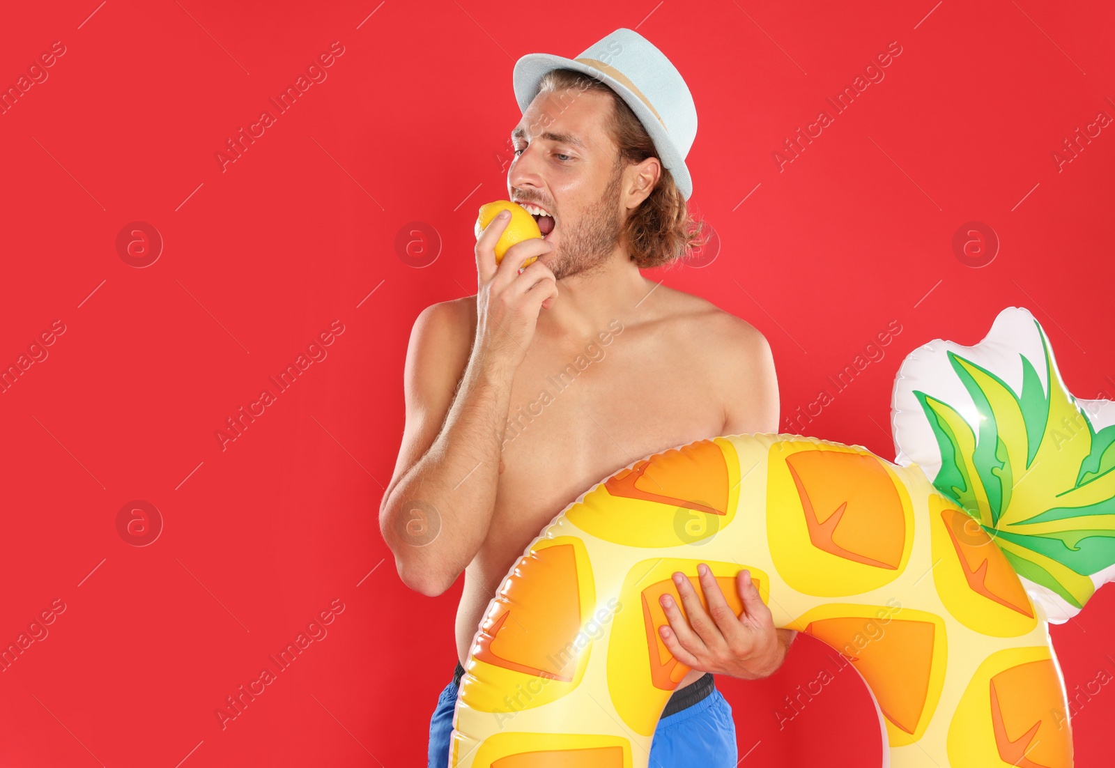 Photo of Attractive young man in swimwear with pineapple inflatable ring eating lemon on red background