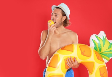 Photo of Attractive young man in swimwear with pineapple inflatable ring eating lemon on red background