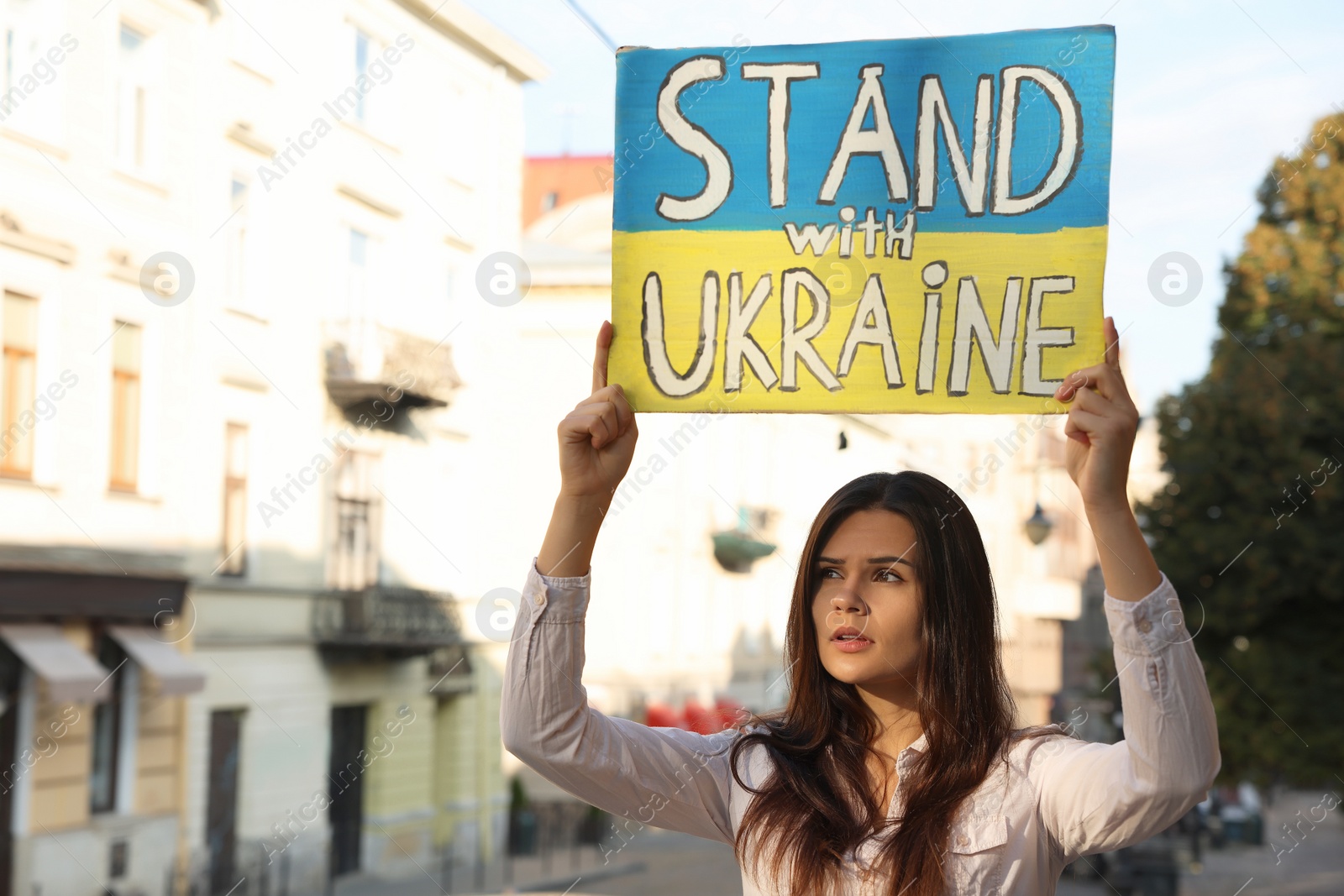 Photo of Sad woman holding poster in colors of national flag and words Stand with Ukraine on city street. Space for text