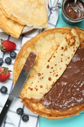 Photo of Tasty crepes with chocolate paste and berries served on turquoise wooden table, flat lay