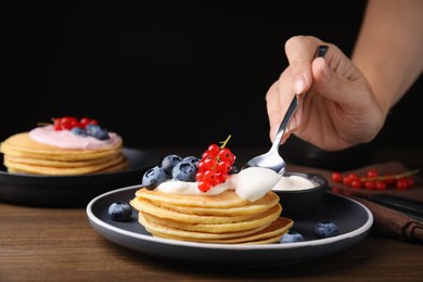 Photo of Woman putting natural yogurt onto tasty pancakes with blueberries and red currants at wooden table, closeup. Space for text
