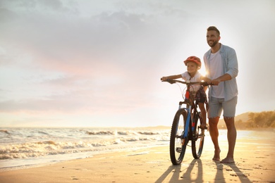Photo of Happy father teaching son to ride bicycle on sandy beach near sea at sunset