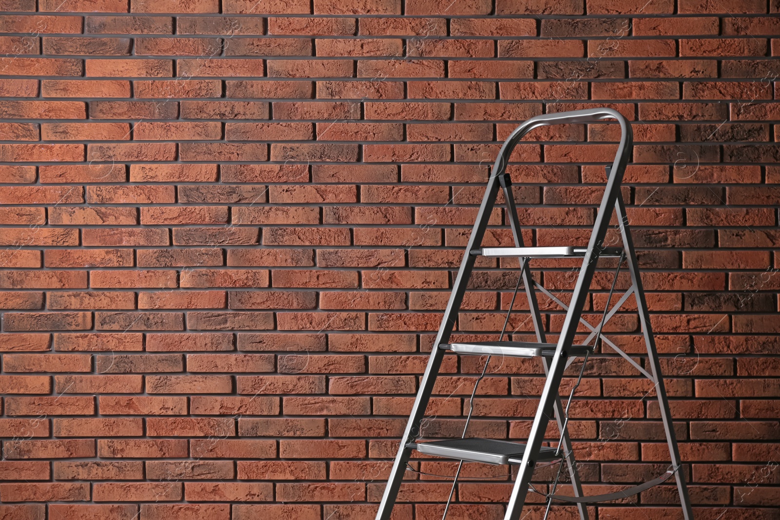 Photo of Modern metal stepladder against brick background. Space for text