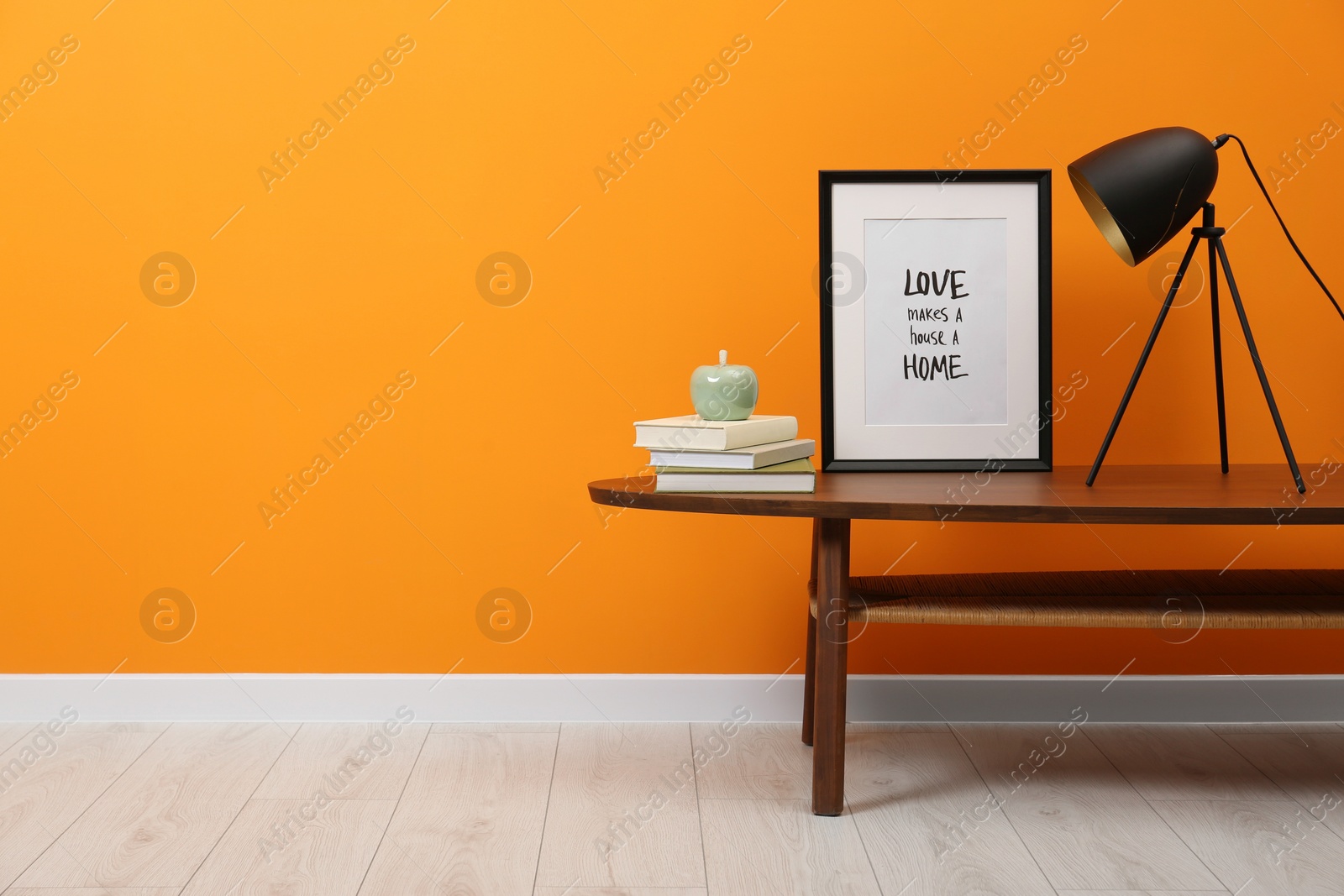 Photo of Lamp, frame, stack of books and ceramic apple on wooden coffee table near orange wall indoors, space for text. Stylish interior design