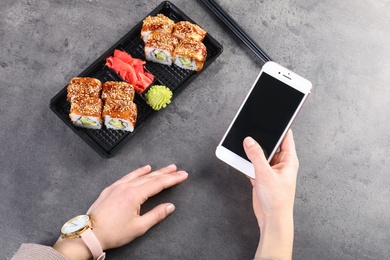 Woman with smartphone and tasty sushi rolls at grey table, space for text. Food delivery