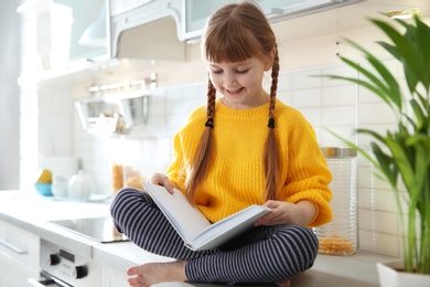 Cute little girl reading book in kitchen at home