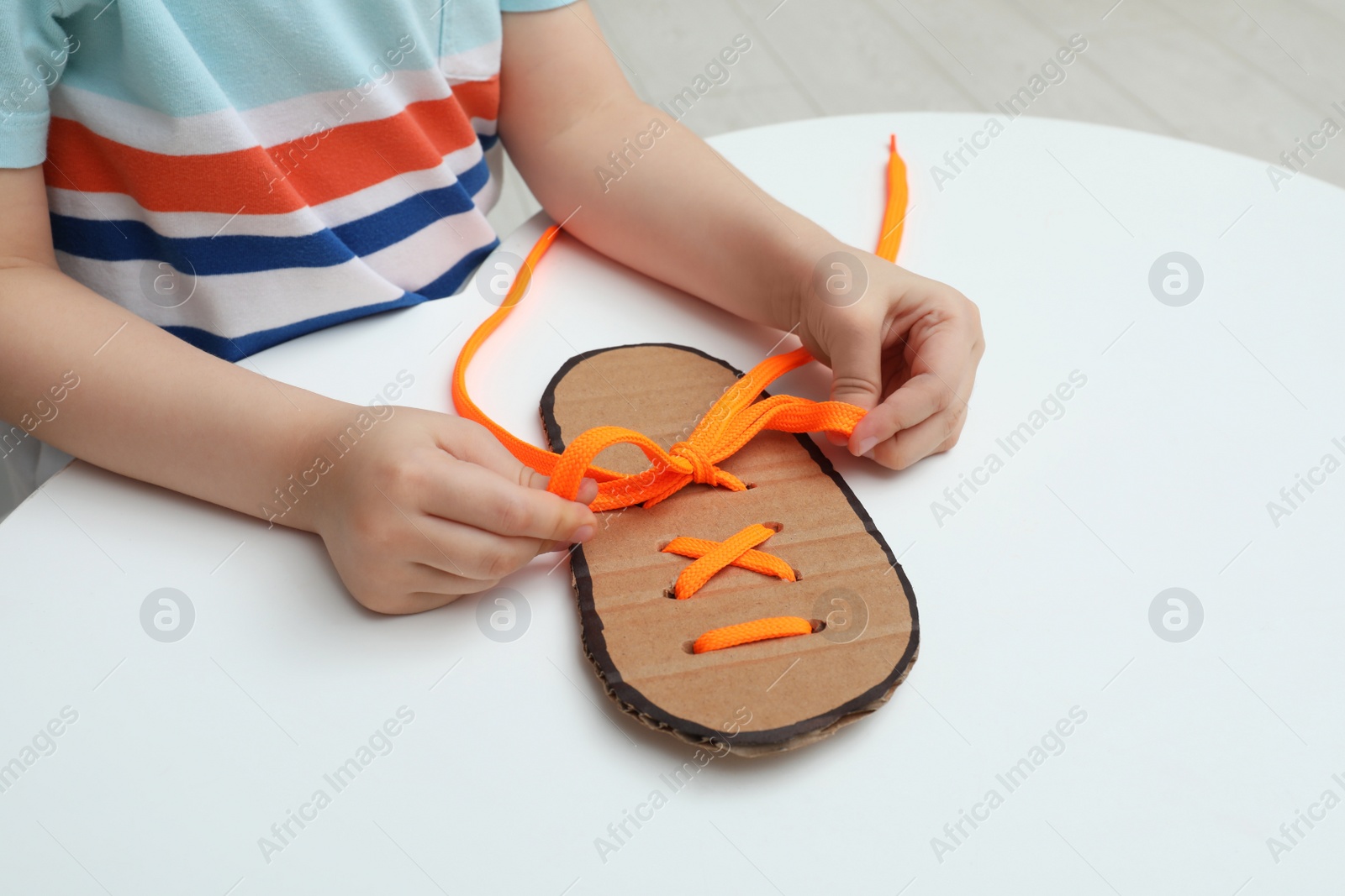 Photo of Little boy tying shoe lace using training cardboard template at white table, closeup