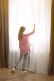 Young woman opening window curtains at home