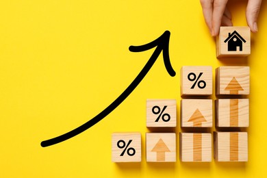 Image of Mortgage rate rising illustrated by upward arrows. Woman putting wooden cube with house icon near other ones on yellow background, top view