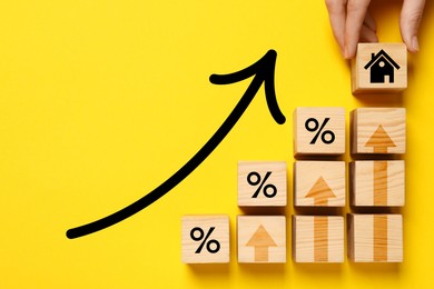 Mortgage rate rising illustrated by upward arrows. Woman putting wooden cube with house icon near other ones on yellow background, top view