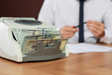 Photo of Modern banknote counter with money and blurred view of man working at wooden table indoors