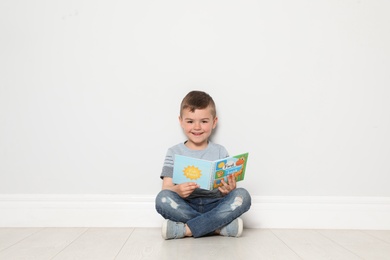 Photo of Cute little boy reading book on floor near white wall, space for text