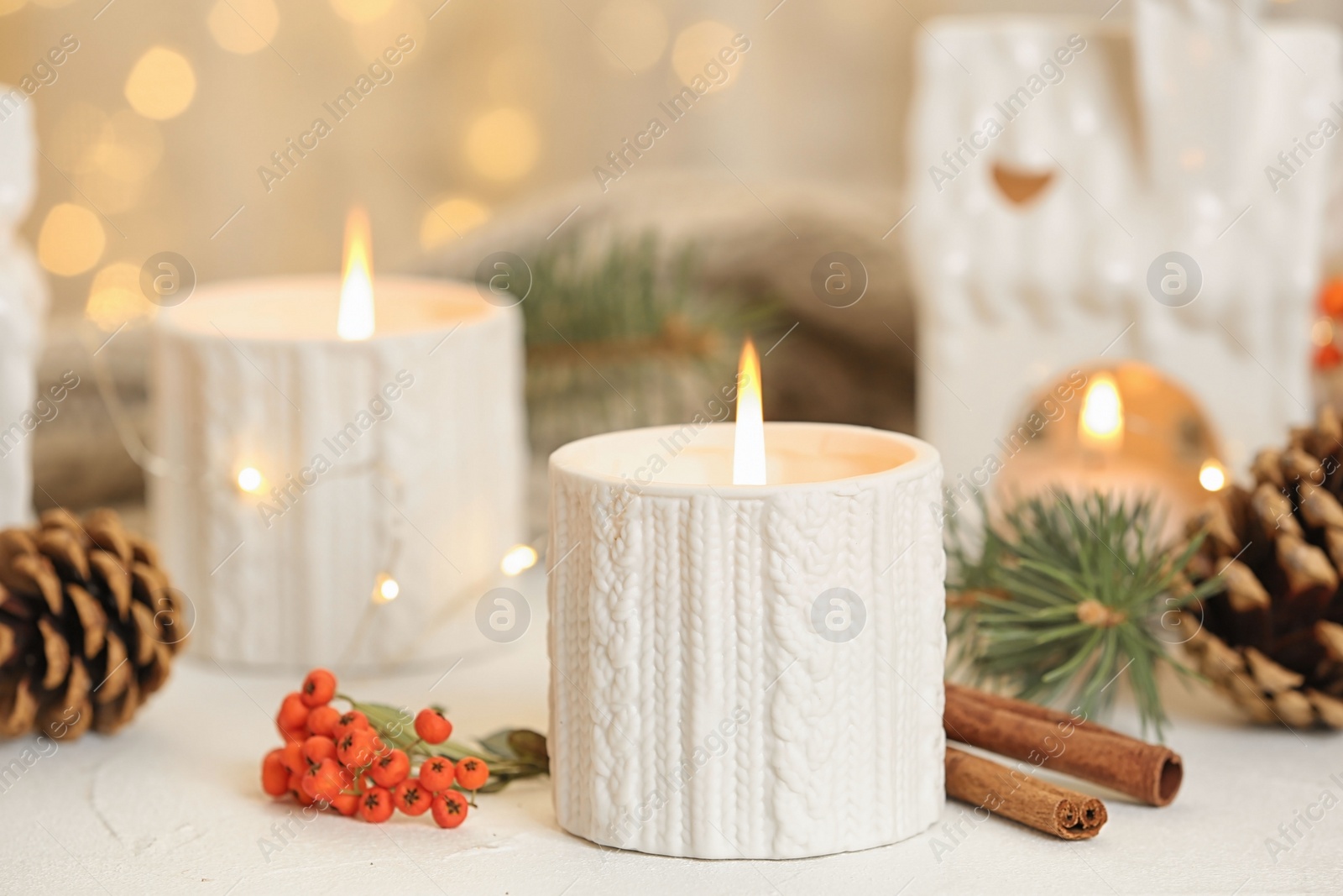 Photo of Holders with burning candles and decoration on white table against blurred Christmas lights