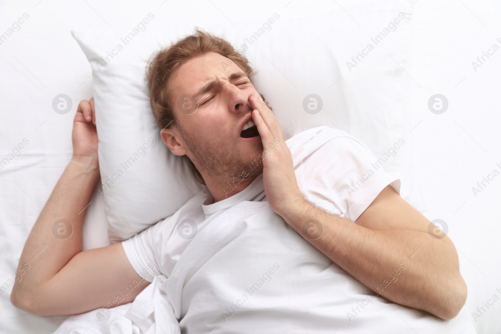 Photo of Handsome young man yawning while lying on pillow in morning, view from above. Bedtime