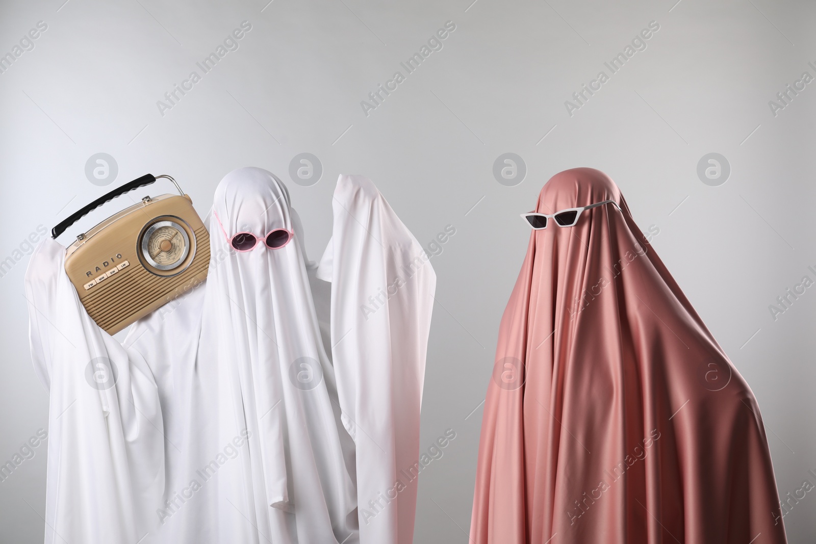 Photo of Glamorous ghosts. Women in color sheets and sunglasses with retro radio receiver on light grey background