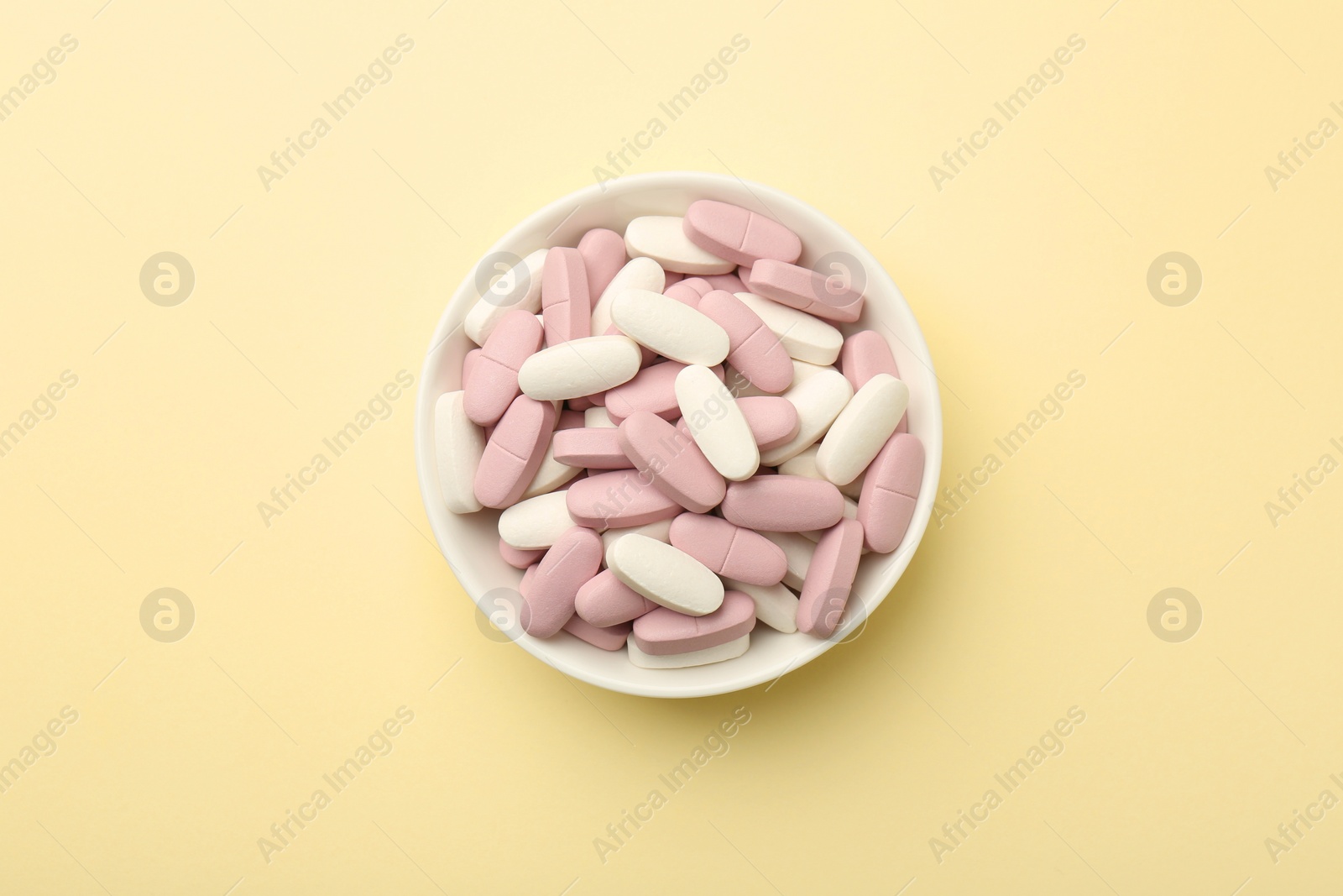 Photo of Different vitamin pills in bowl on pale yellow background, top view