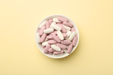 Photo of Different vitamin pills in bowl on pale yellow background, top view