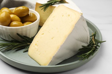 Photo of Plate with pieces of tasty camembert cheese, olives and rosemary on white table, closeup