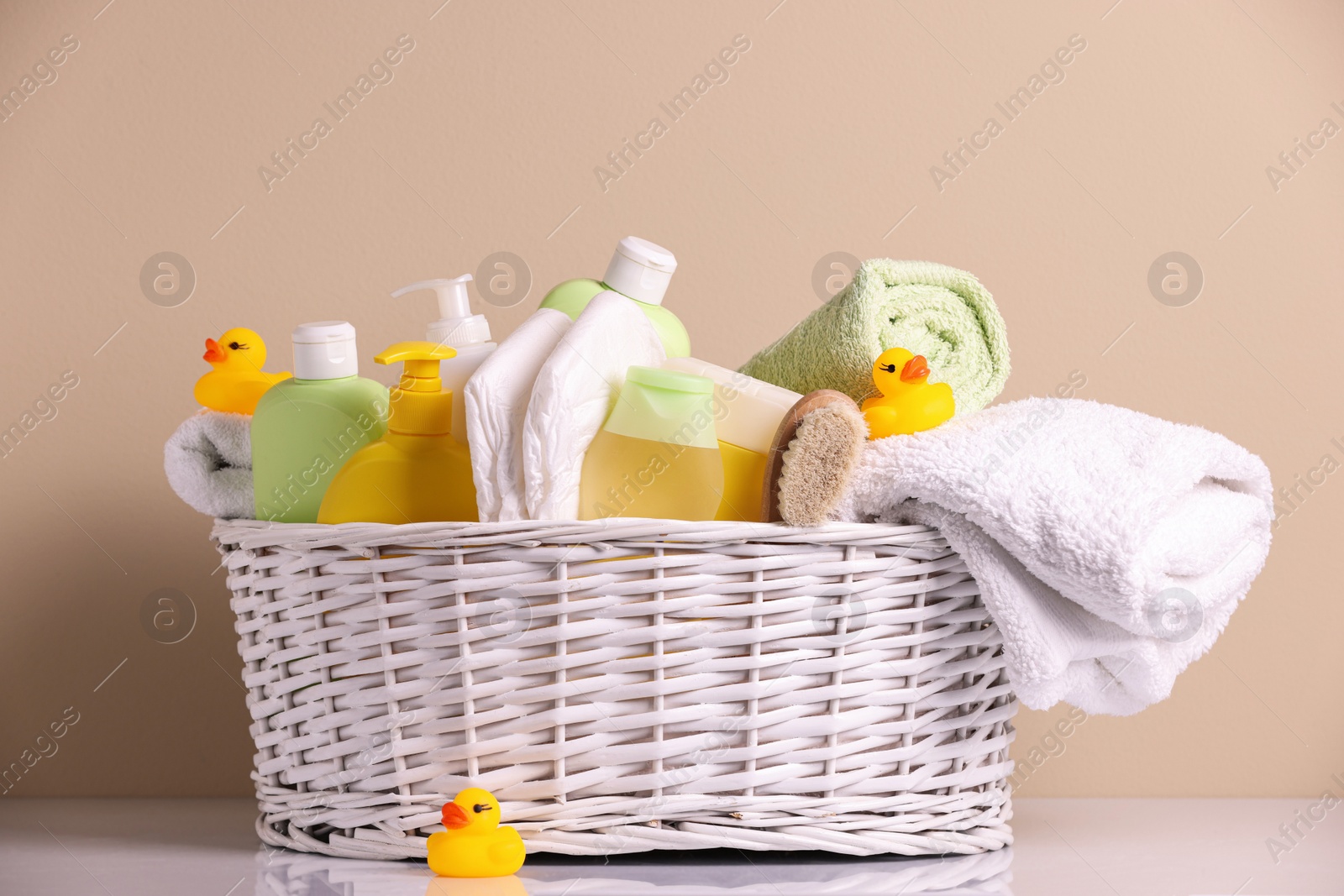 Photo of Wicker basket with baby cosmetic products, bath accessories and rubber ducks on white table against beige background