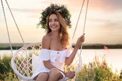 Photo of Young woman wearing wreath made of beautiful flowers on swing chair outdoors