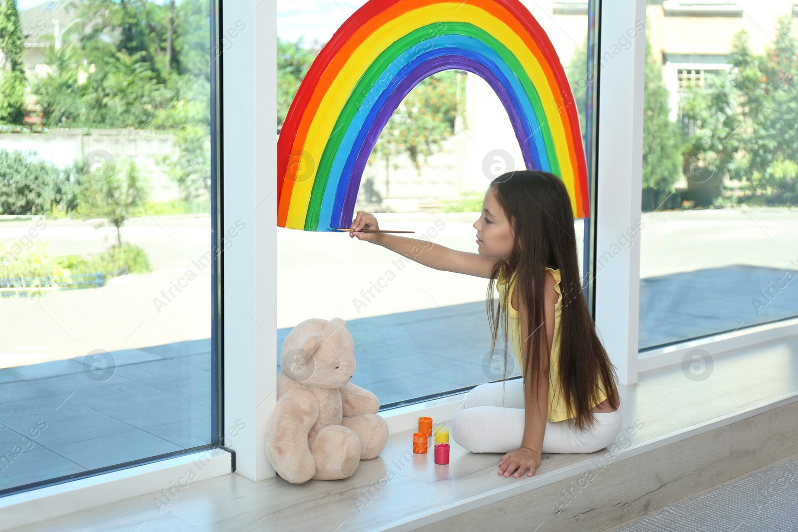 Photo of Little girl drawing rainbow on window with paints indoors. Stay at home concept