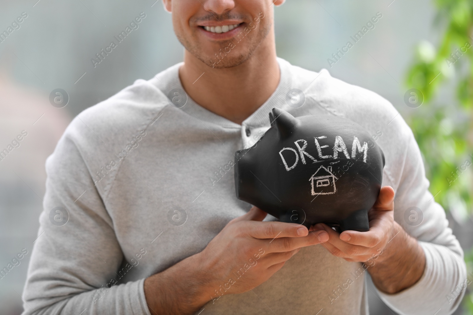 Photo of Man holding piggy bank with word DREAM against blurred background, closeup