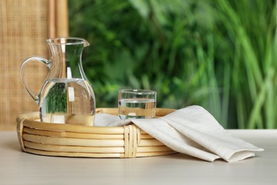 Photo of Stylish wicker tray with jug and glass of water on white table against blurred background