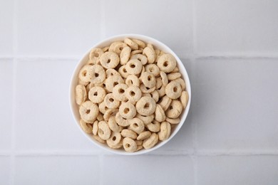 Tasty cereal rings in bowl on white tiled table, top view