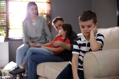 Photo of Unhappy little boy feeling jealous while parents spending time with his sister at home