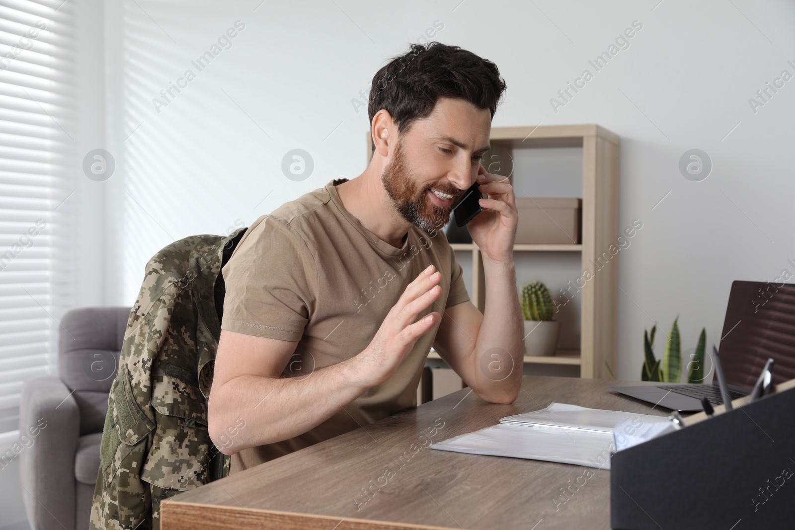 Photo of Happy soldier talking on phone at wooden table indoors. Military service