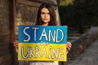 Photo of Sad woman holding poster in colors of national flag and words Stand with Ukraine on city street