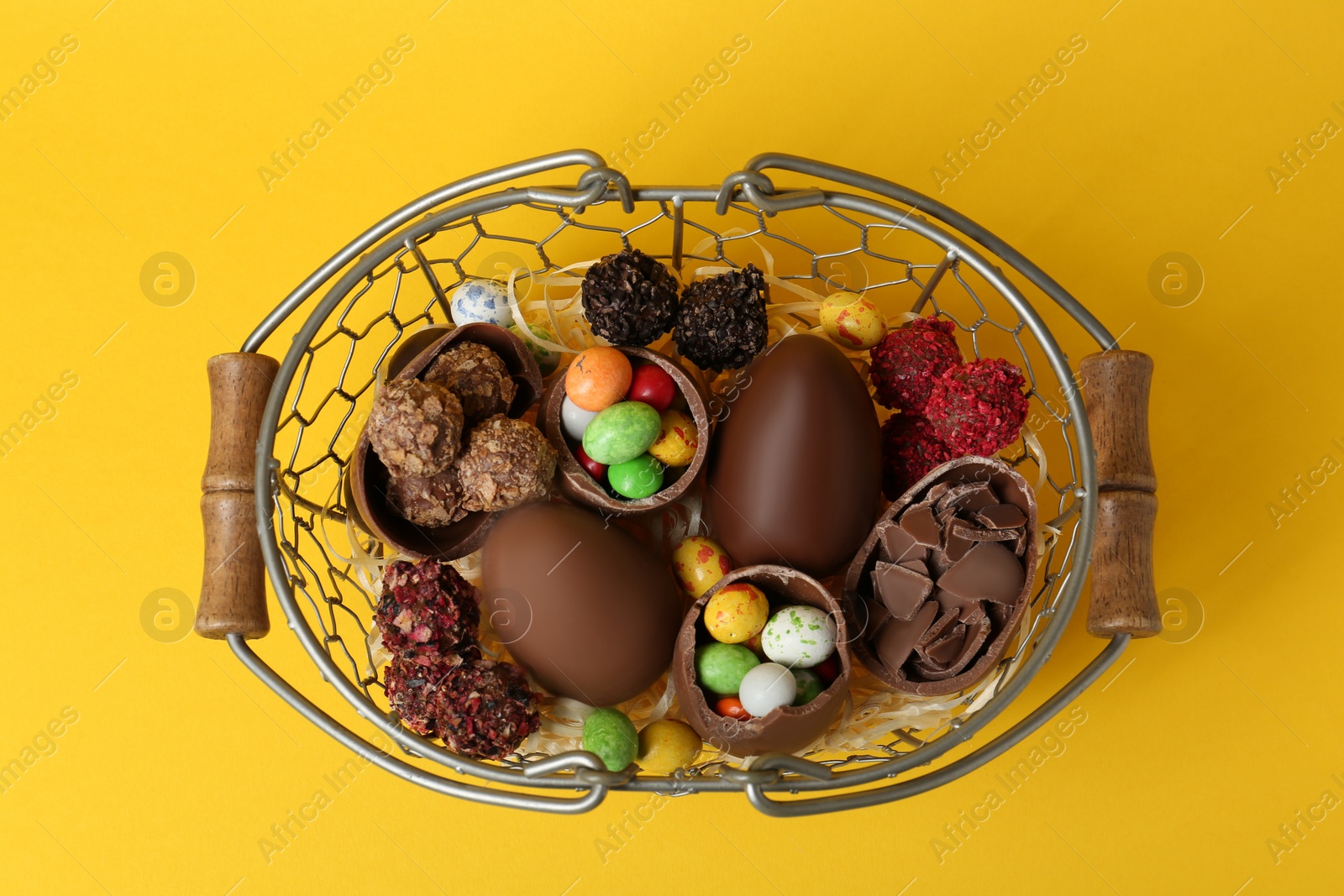 Photo of Basket with chocolate eggs and candies on yellow background, top view