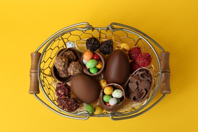 Basket with chocolate eggs and candies on yellow background, top view