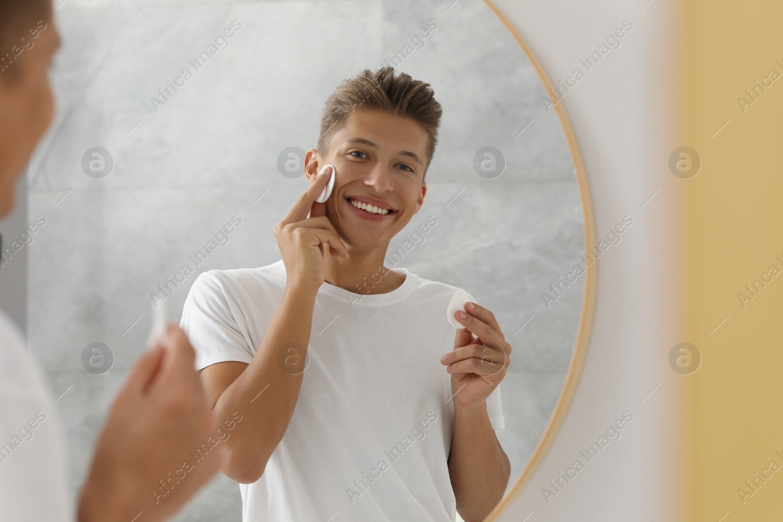 Photo of Handsome young man cleaning face with cotton pad near mirror in bathroom