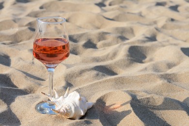 Glass of tasty rose wine and seashell on sand, space for text
