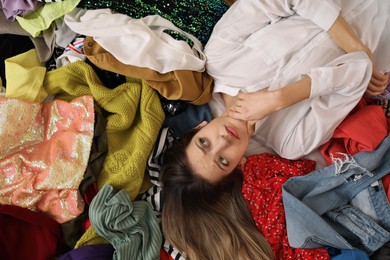 Photo of Young woman lying on pile of different clothes, top view. Fast fashion concept