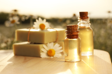 Bottles of chamomile essential oil and soap bars on wooden table in field