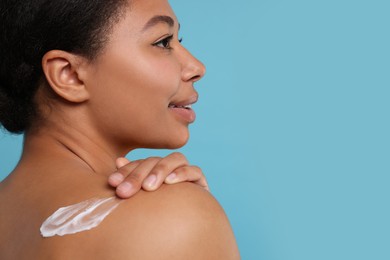 Young woman applying body cream onto back on light blue background. Space for text
