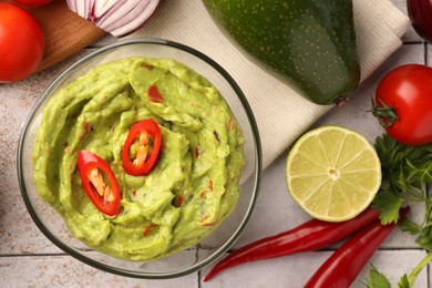 Bowl of delicious guacamole and ingredients on white tiled table, flat lay