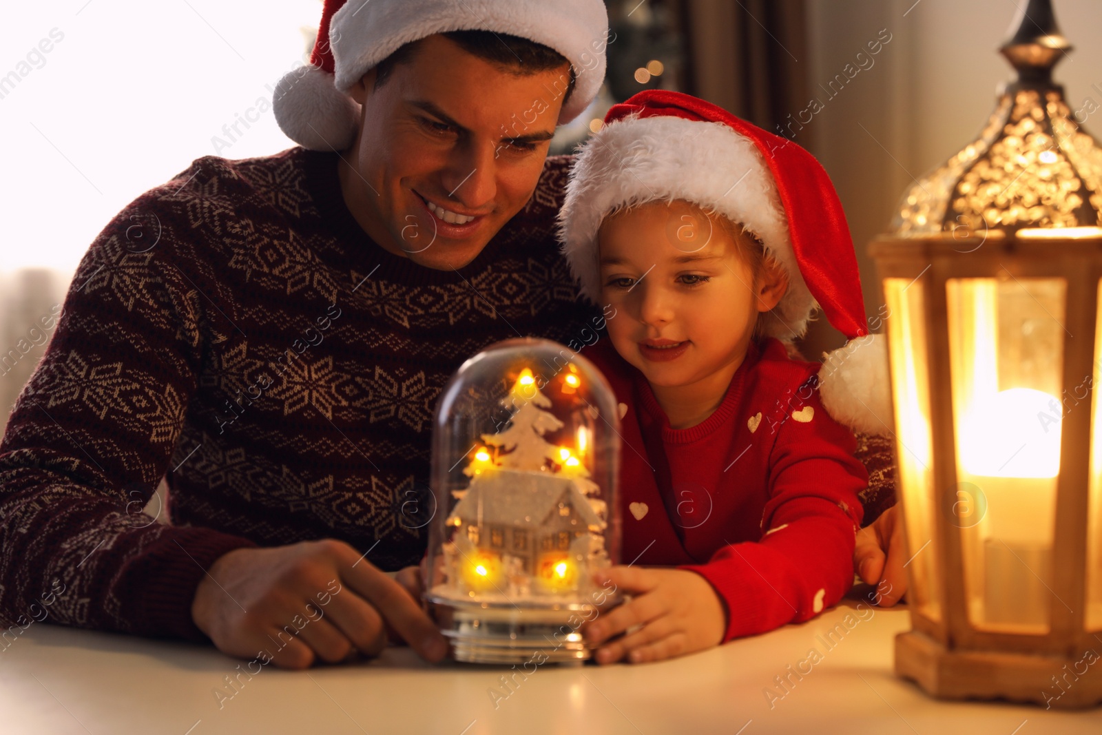 Photo of Father and daughter in Santa hats playing with snow globe at table