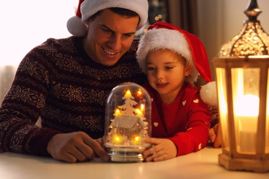 Father and daughter in Santa hats playing with snow globe at table