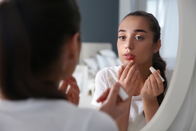 Photo of Young woman with herpes applying lip balm in front of mirror at home