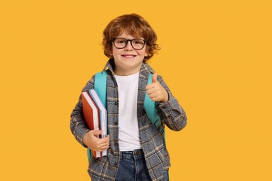 Photo of Happy schoolboy with backpack and books showing thumb up gesture on orange background
