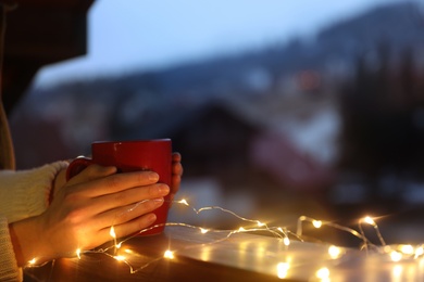 Photo of Woman holding cup of hot beverage on balcony decorated with Christmas lights, closeup with space for text. Winter evening