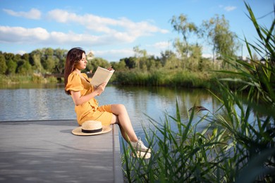 Young woman reading book near lake on sunny day