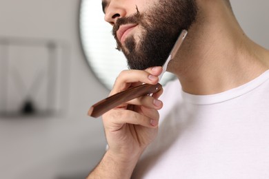 Photo of Handsome young man shaving beard with blade indoors, closeup